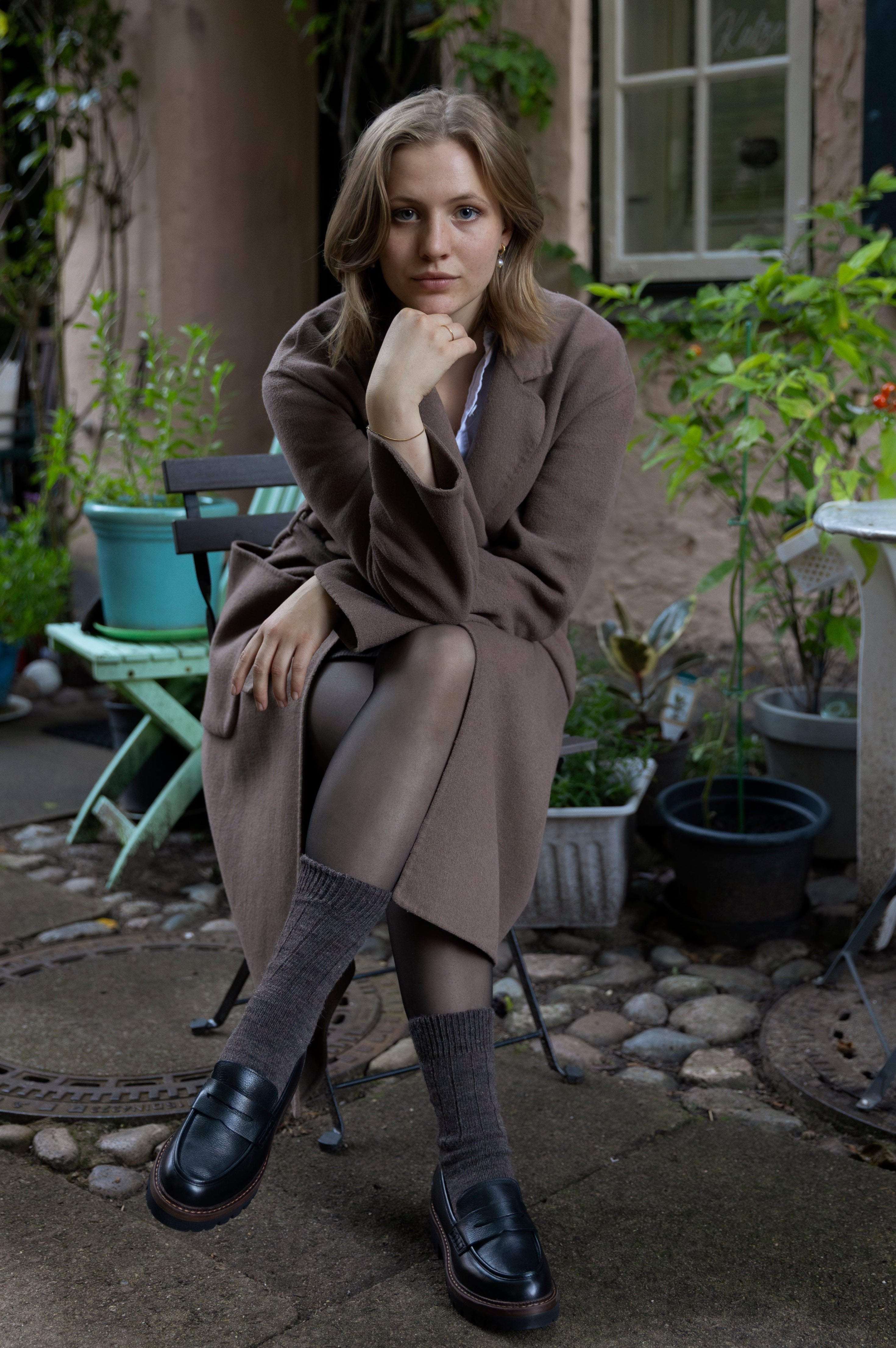 Woman sitting outdoors in a garden setting with plants and a building in the background
