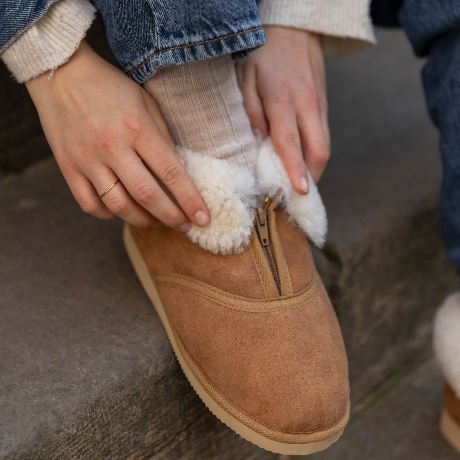 Person wearing brown suede shoes with white fuzzy lining, sitting on a stone surface.
