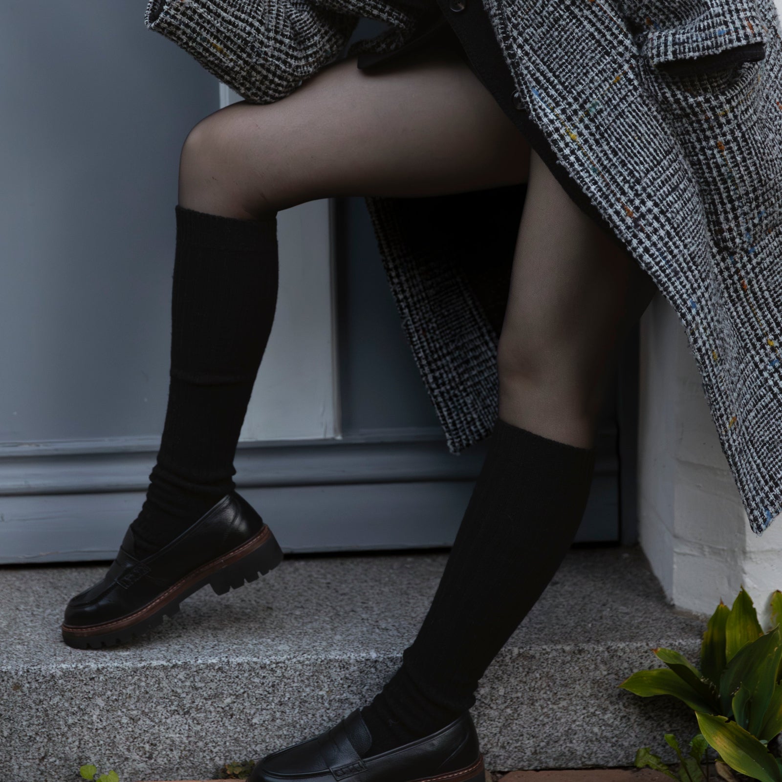 Woman wearing a long gray coat leaning against a building.