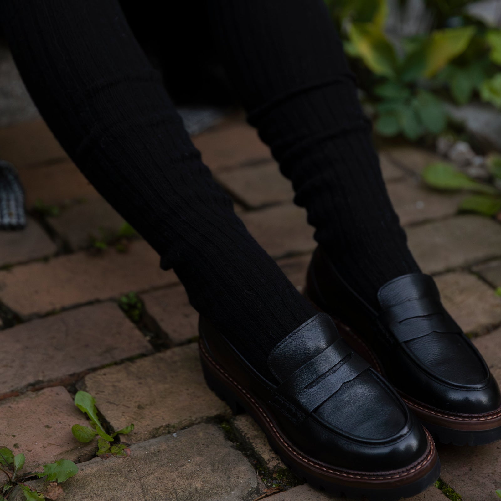 Person wearing black knee-high sock and black tights sitting on a brick surface.