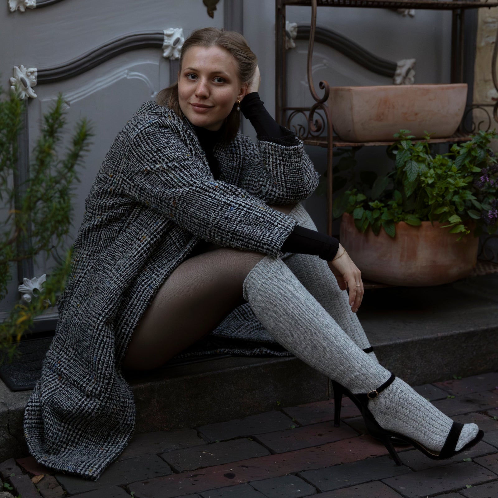 Woman sitting on steps wearing a gray coat and knee-high socks with black shoes.