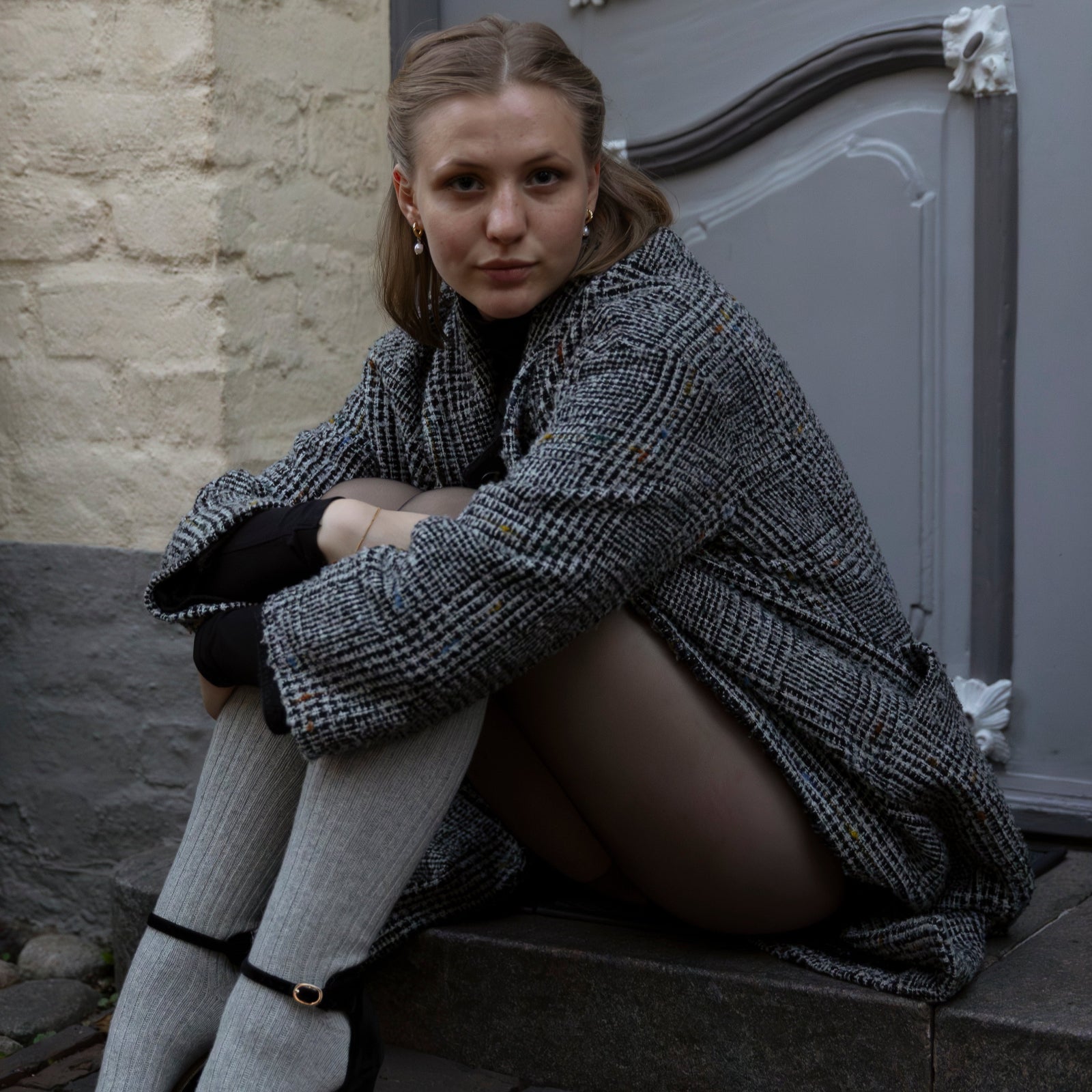 Woman sitting on steps wearing a coat and high heels, with a stone wall and door in the background.